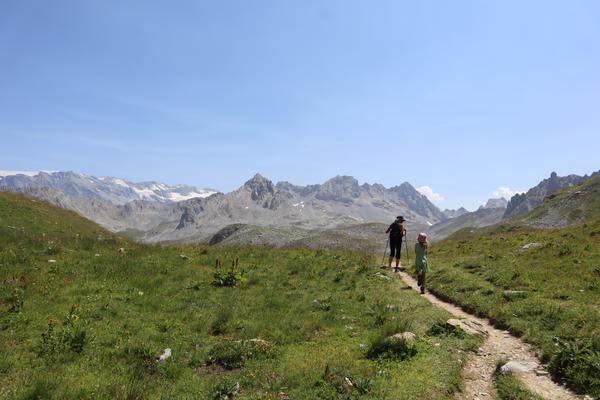 Two hikers, one adult and one child, walk along a winding dirt path through a vibrant green alpine meadow